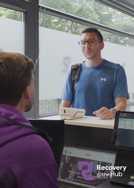 A person in a blue athletic shirt stands at a reception desk, engaging with another individual seated at a computer.
