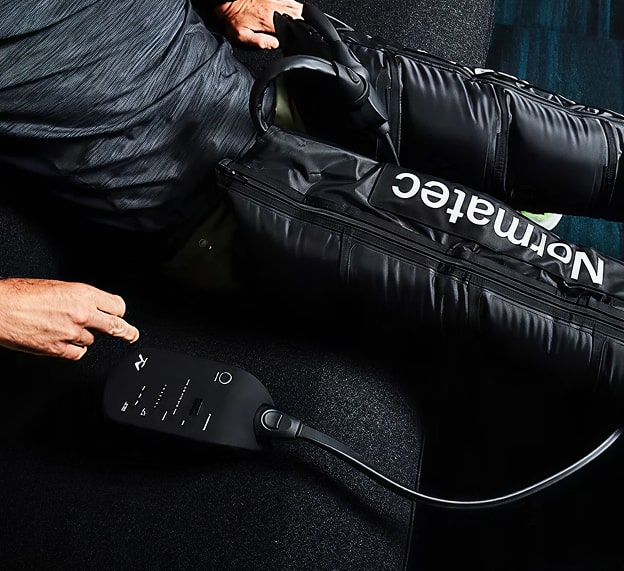 A person uses a black Normatec compression device on their legs while seated, with a control unit positioned nearby.