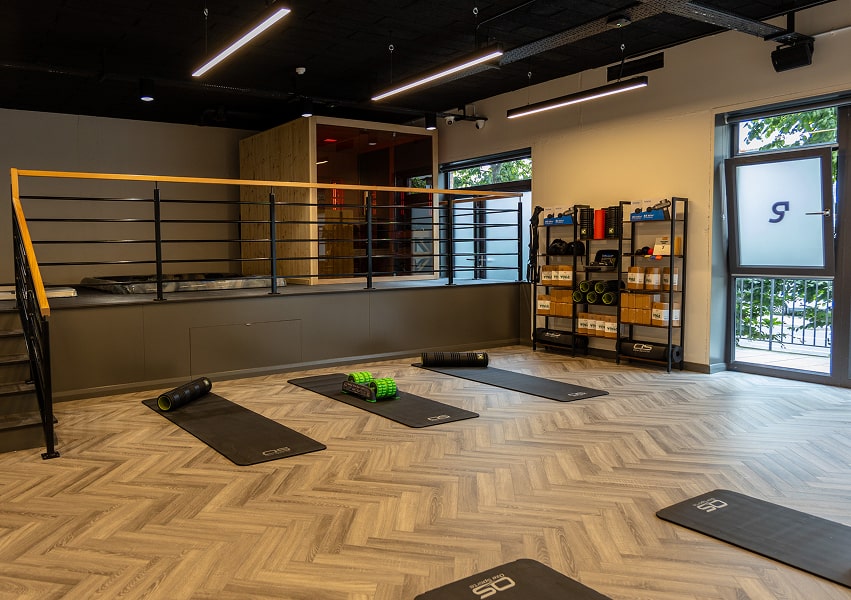 A modern fitness studio with exercise mats, foam rollers, and shelves of equipment, featuring large windows and a stylish design.