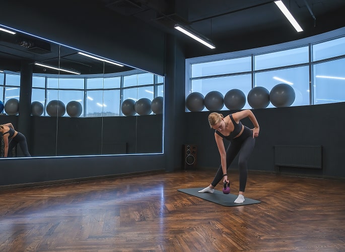 A woman in athletic wear performs a dumbbell exercise on a mat in a modern gym, reflecting in a large mirror with fitness balls in the background.