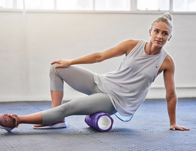 A person demonstrating a foam rolling exercise on a purple foam roller in a bright, minimal gym space.