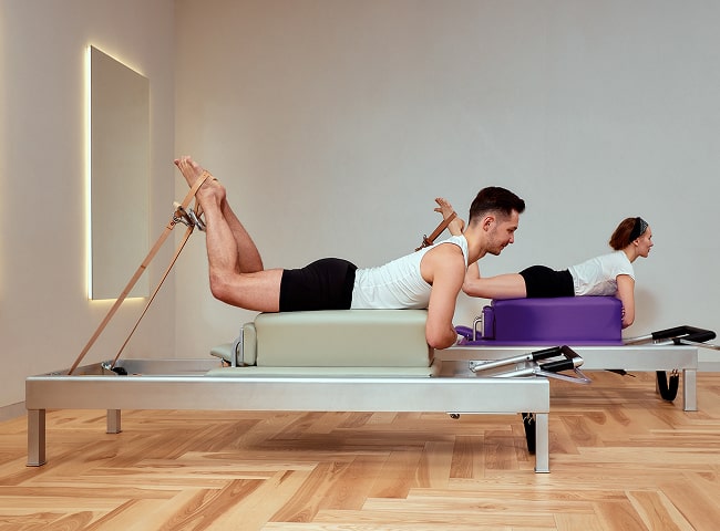 Two individuals are practicing pilates on reformer machines in a bright, minimalist studio with wooden flooring.