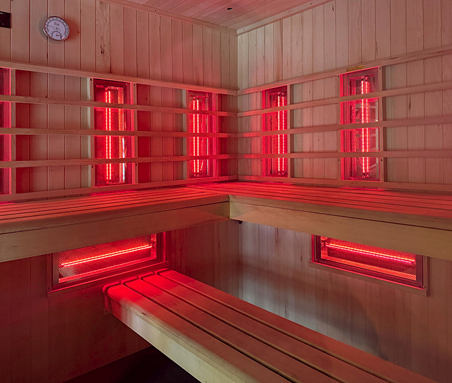 Interior of a red-lit sauna featuring wooden benches, glass doors, and glowing wall panels, creating a warm and relaxing ambiance.